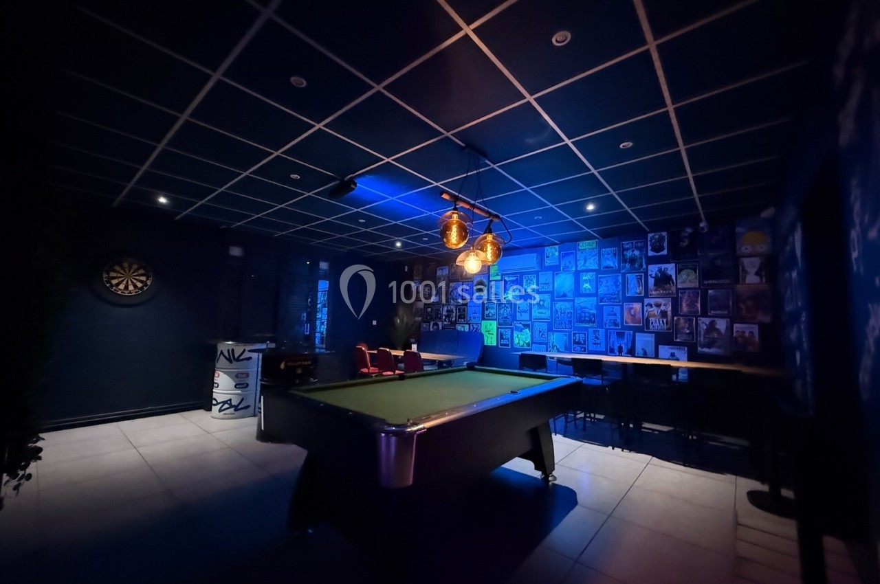 Salle sombre avec une table de billard éclairée, un mur décoré de photos et une cible de fléchettes au fond.