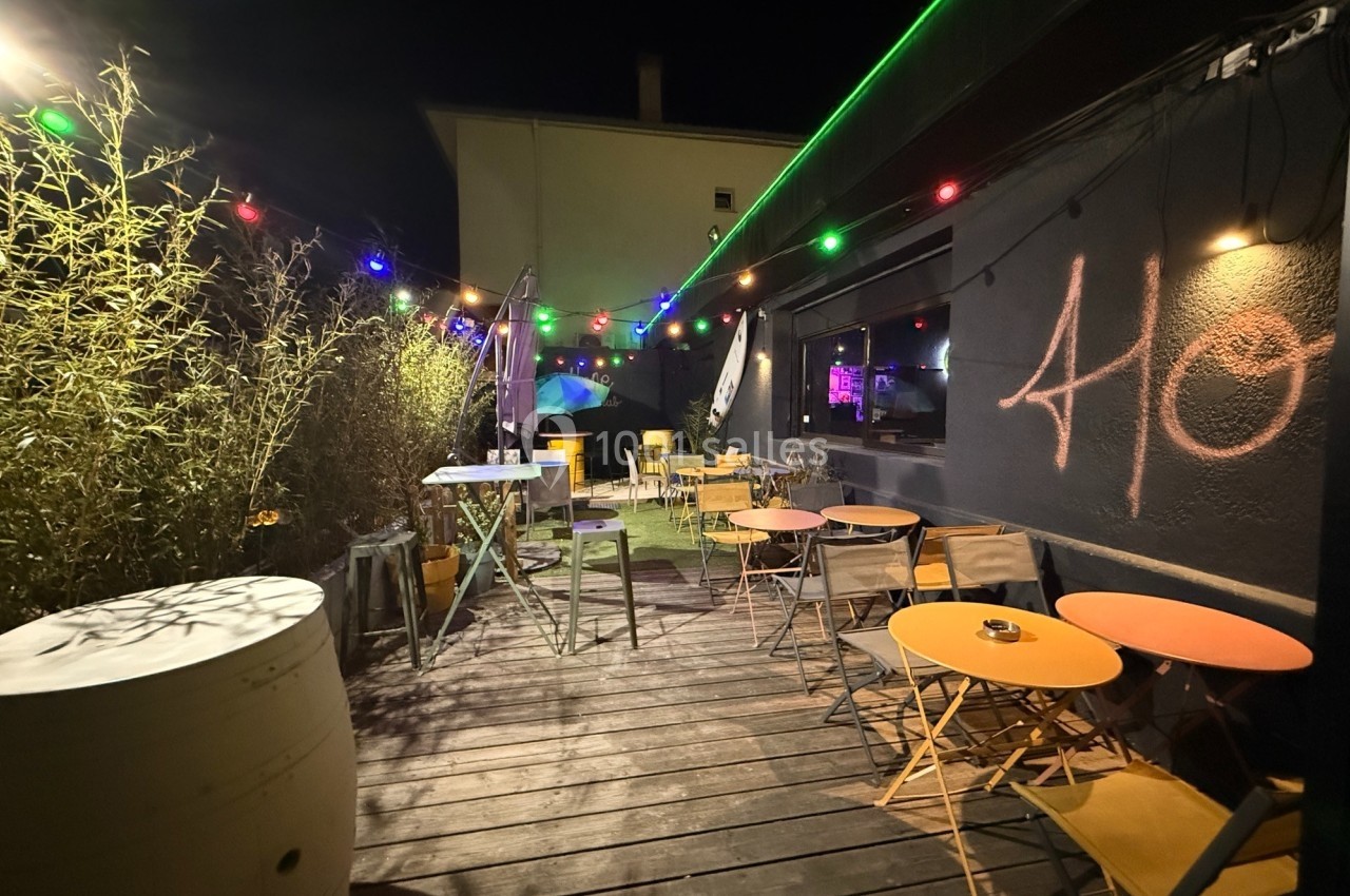 Terrasse extérieure en bois éclairée par des guirlandes lumineuses colorées, avec tables et chaises disposées.