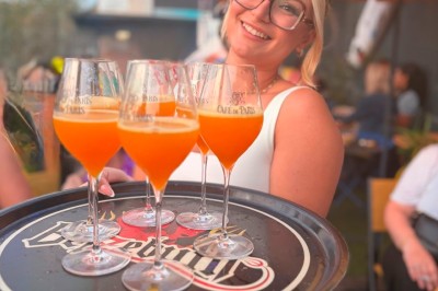 Miniature Location salle Mérignac (Gironde) - Hope Bar #8 Serveuse souriante tenant un plateau avec quatre cocktails orange sur une terrasse animée et colorée.