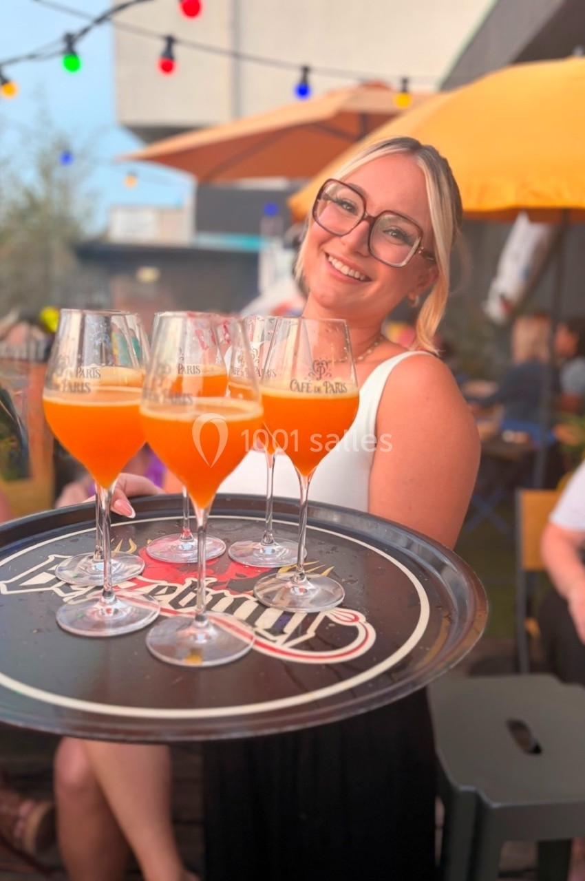 Serveuse souriante tenant un plateau avec quatre cocktails orange sur une terrasse animée et colorée.