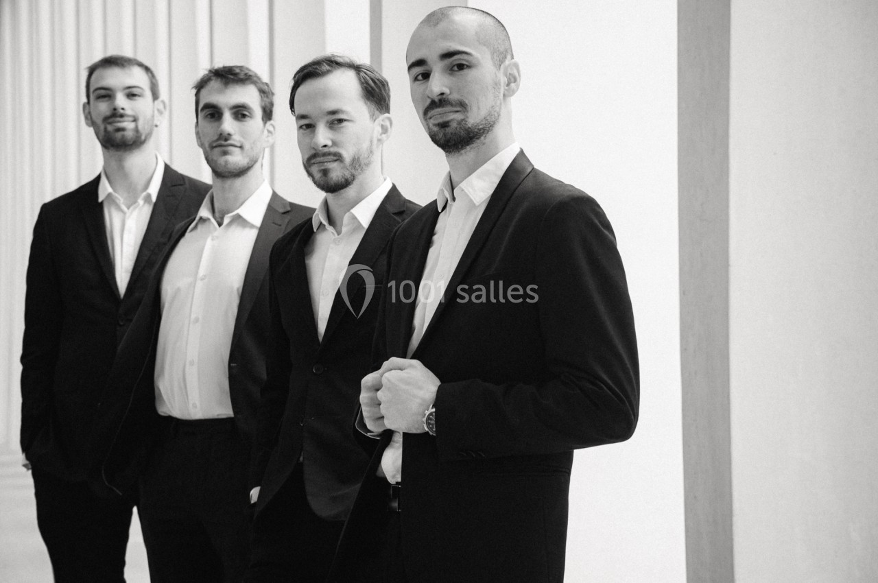 Quatre hommes en costume posent debout dans un espace lumineux avec des colonnes en arrière-plan.