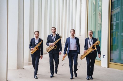 Miniature DJ Saint-Gilles (Bruxelles-Capitale) - 441 Quartet #3 Quatre musiciens en costume saluent sur une scène éclairée, entourés de bougies, dans un espace intérieur industriel.