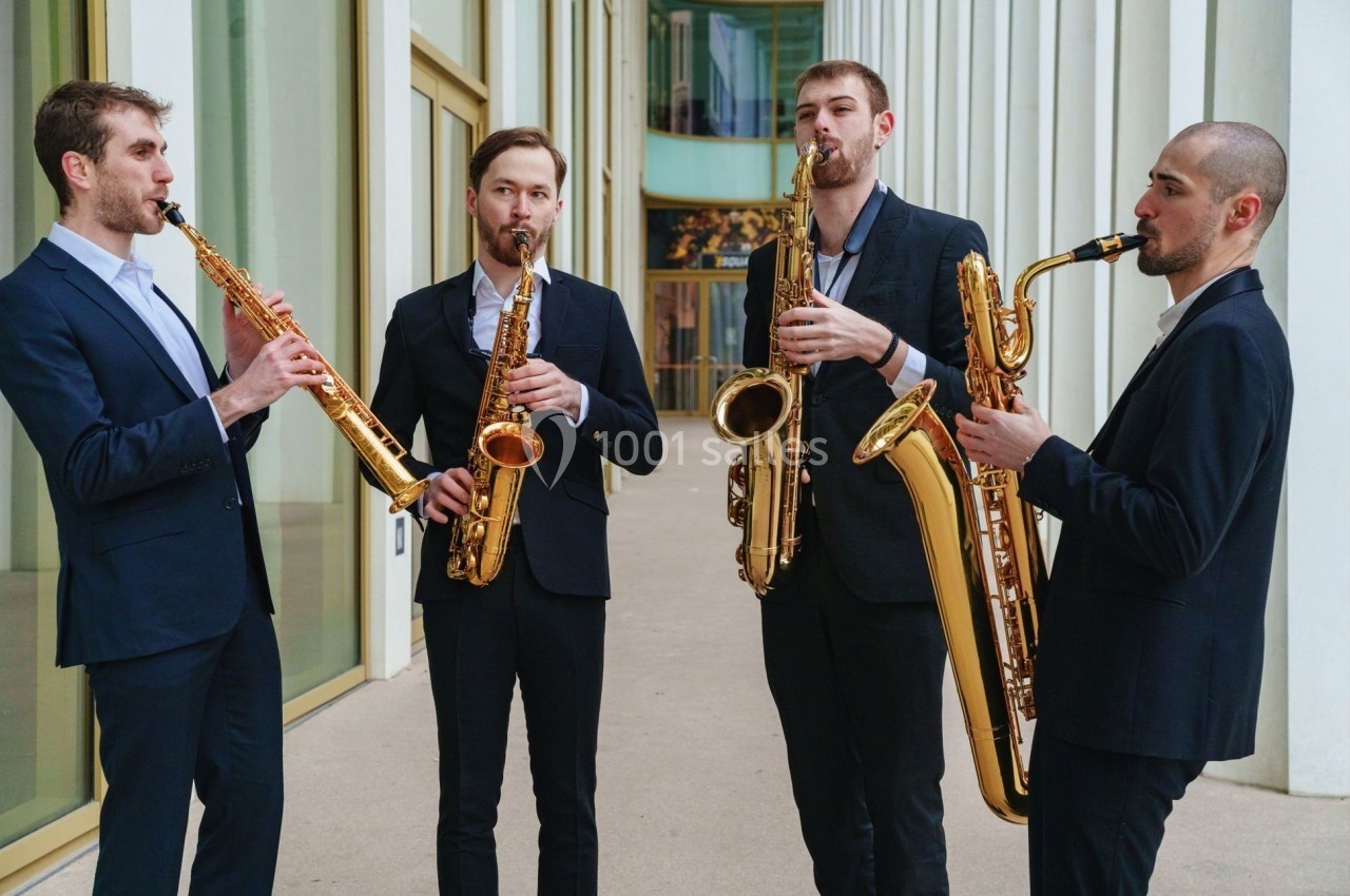 Quatre musiciens en costumes jouent du saxophone devant un bâtiment moderne avec de grandes fenêtres.