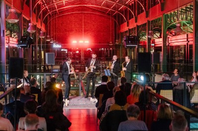 Miniature DJ Saint-Gilles (Bruxelles-Capitale) - 441 Quartet #2 Quatre musiciens en costume saluent sur une scène éclairée, entourés de bougies, dans un espace intérieur industriel.