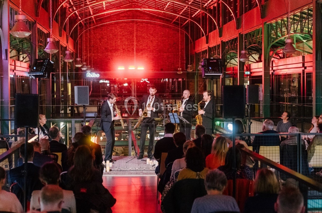 Un groupe de musiciens joue sur une scène dans un espace intérieur éclairé de lumières rouges, devant un public assis.