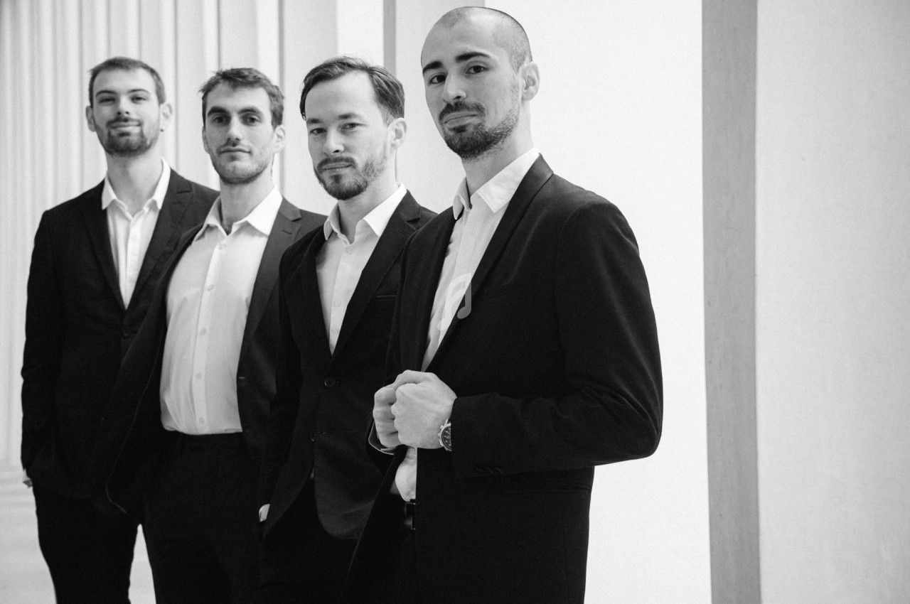 Quatre hommes en costume posant debout dans un espace lumineux avec des colonnes en arrière-plan.