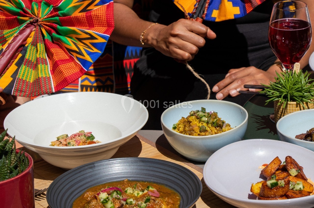Des plats colorés et variés disposés sur une table, avec des mains tenant un éventail et un verre de boisson rouge.