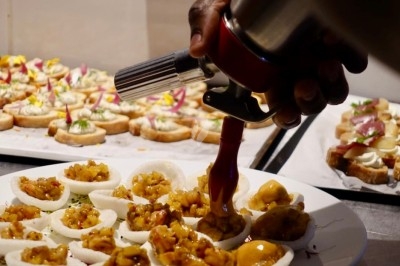 Une main utilise un siphon pour garnir des bouchées apéritives sur une assiette dans une cuisine.