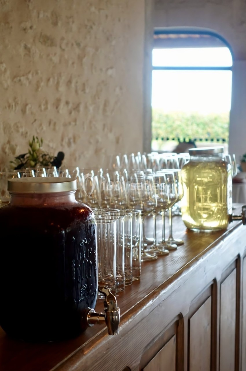Deux jarres de boissons avec des verres alignés sur un comptoir en bois dans une pièce lumineuse.