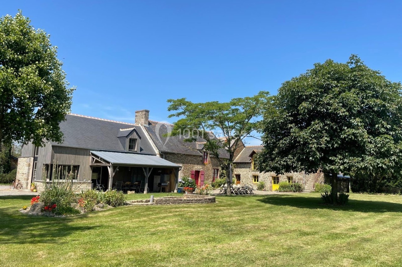 Maison en pierre avec toit en ardoise, entourée d'un grand jardin arboré sous un ciel bleu.