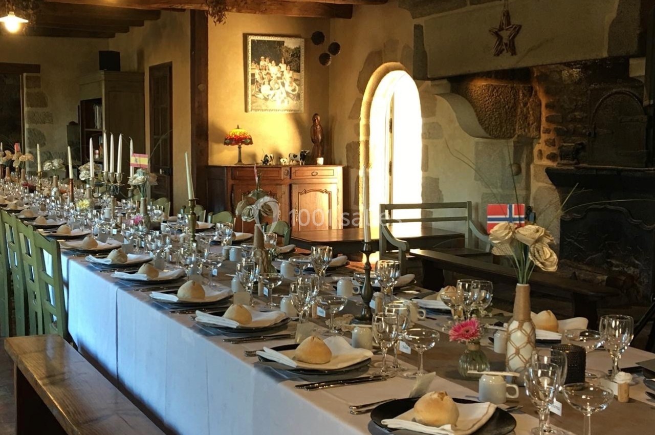 Grande table dressée pour un repas festif dans une salle rustique avec poutres apparentes et cheminée en pierre.