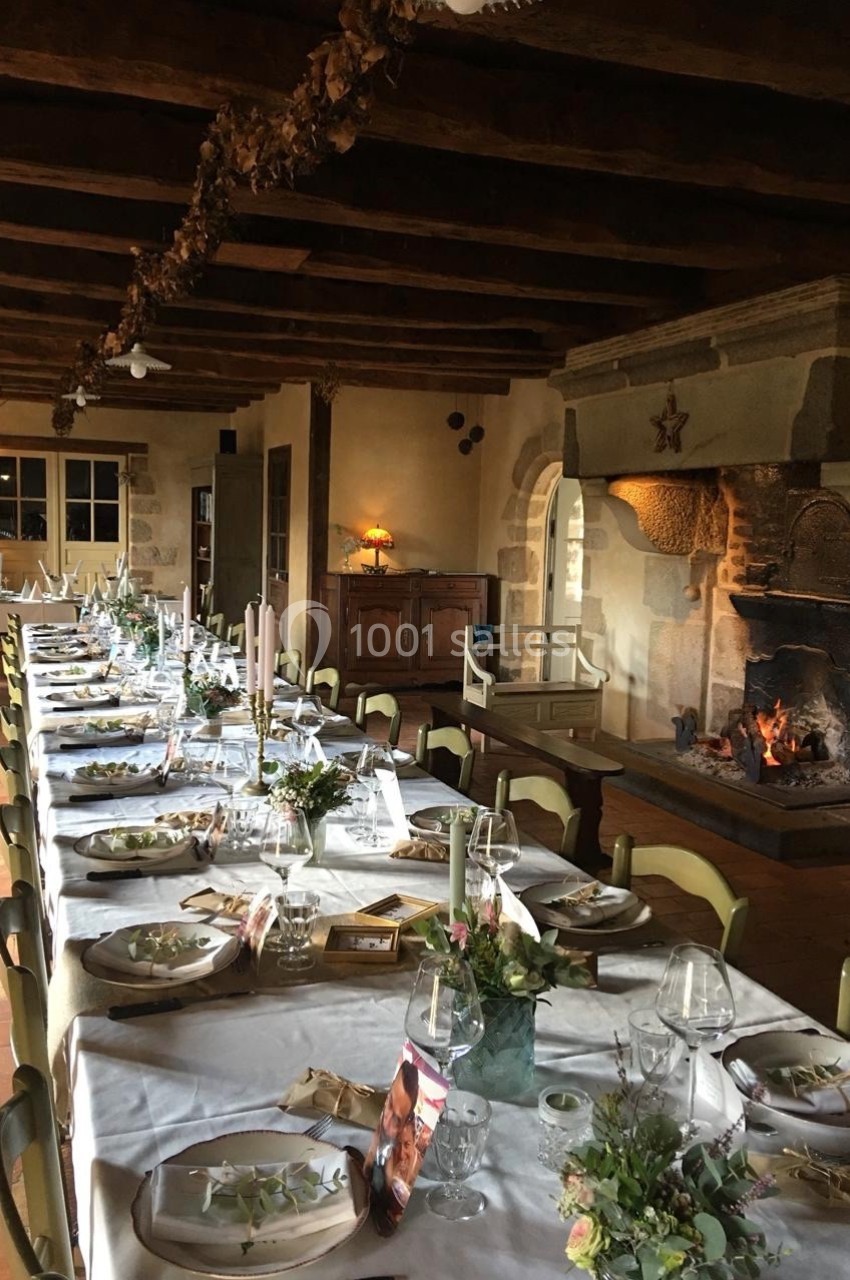 Grande table dressée avec des décorations florales dans une salle rustique avec cheminée en pierre allumée.