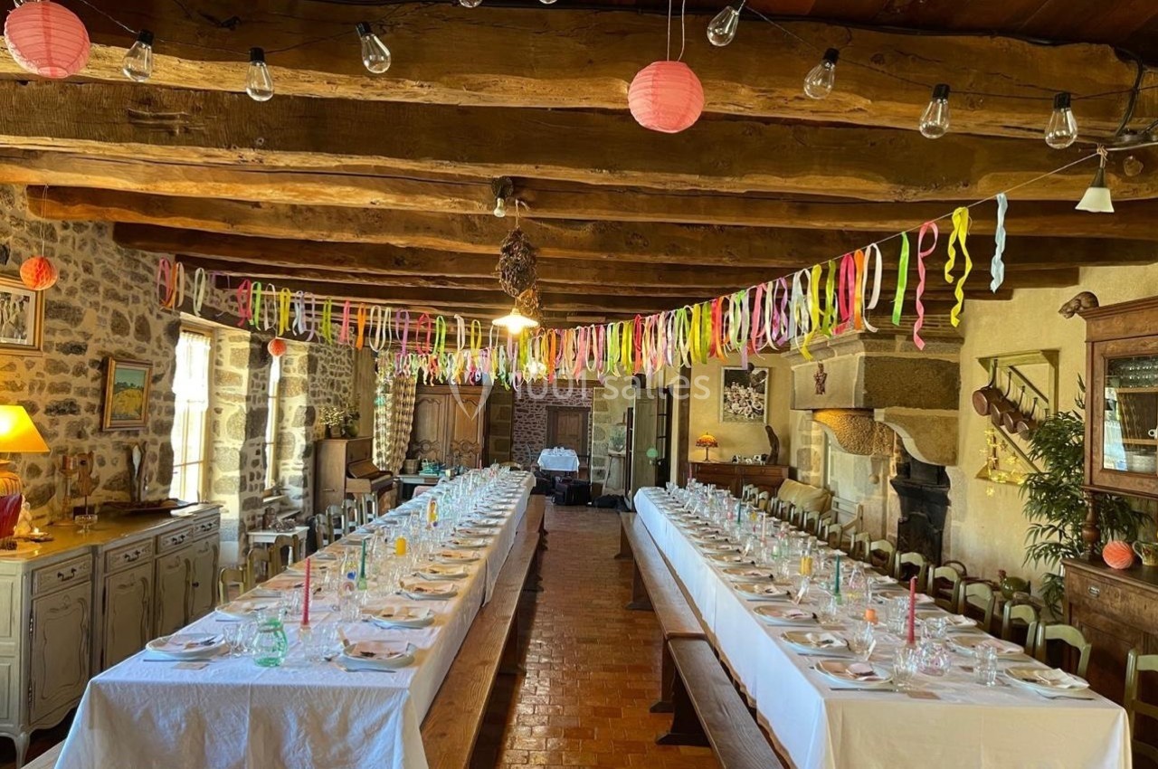 Salle rustique décorée pour un événement, avec deux longues tables dressées, guirlandes colorées et poutres apparentes.
