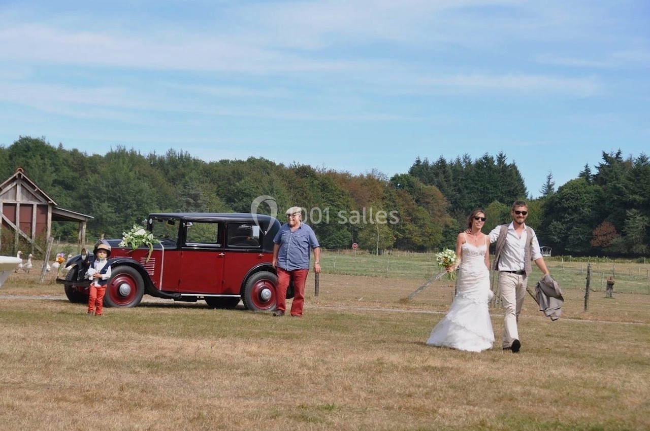 Un couple en tenue de mariage marche dans un champ, suivi d'un enfant, d'un homme et d'une voiture ancienne rouge.