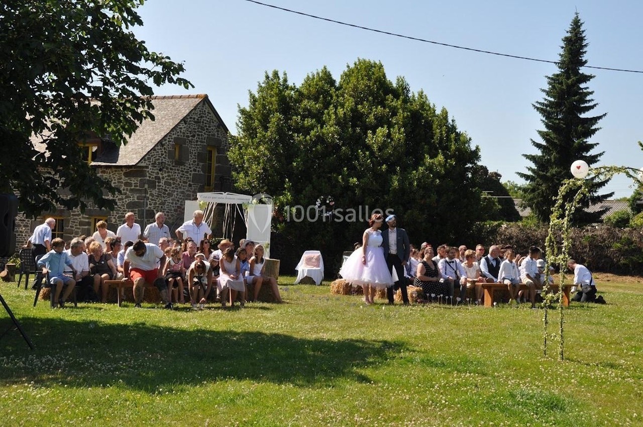 Cérémonie en plein air avec des invités assis sur des bottes de paille, devant une maison en pierre et des arbres.