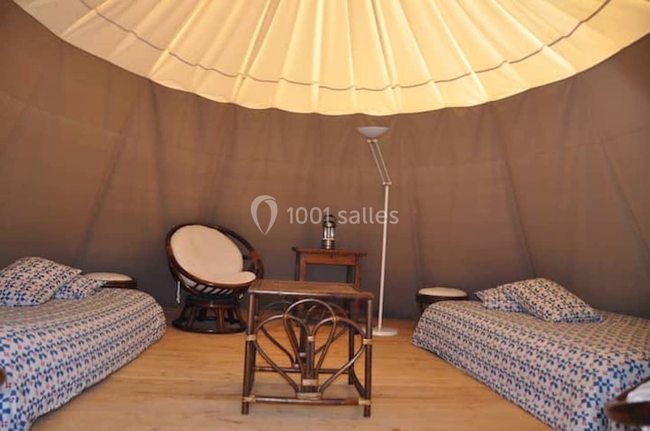 Intérieur d'une tente aménagée avec deux lits, un fauteuil, une table basse et une lampe sur un sol en bois.