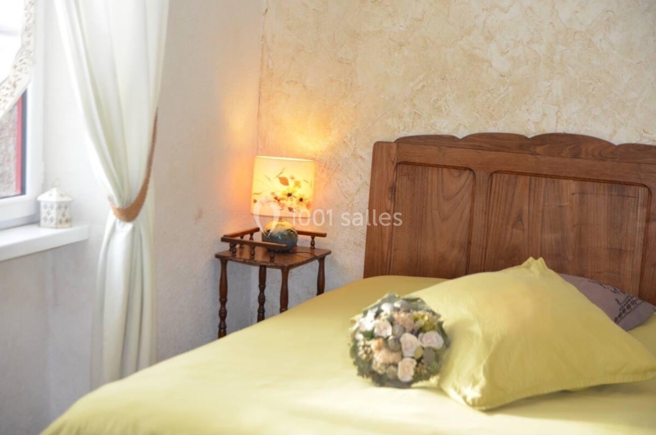 Lit avec draps jaunes, bouquet de fleurs posé dessus, lampe allumée sur une table en bois près d'une fenêtre avec rideaux.