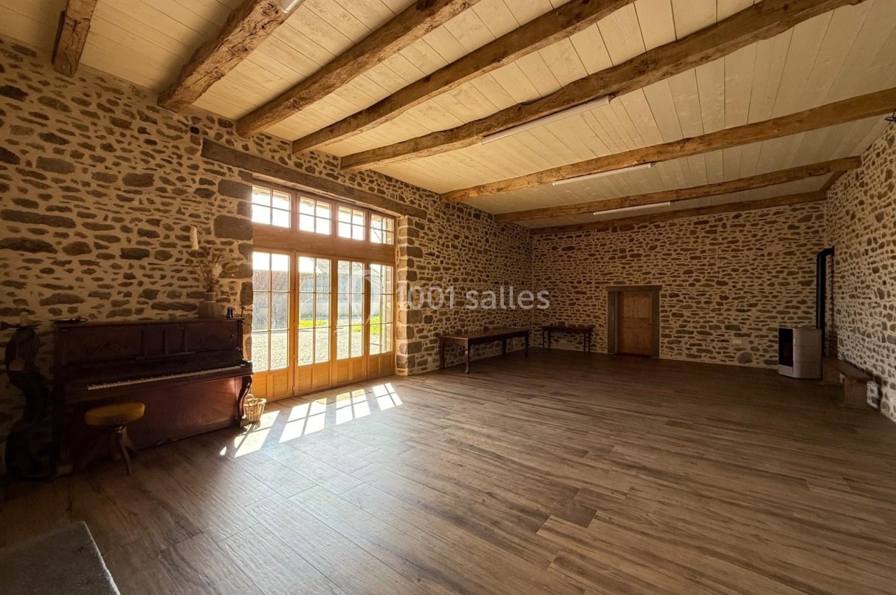 Salle lumineuse avec murs en pierre, parquet en bois, poutres apparentes et un piano près d'une grande baie vitrée.
