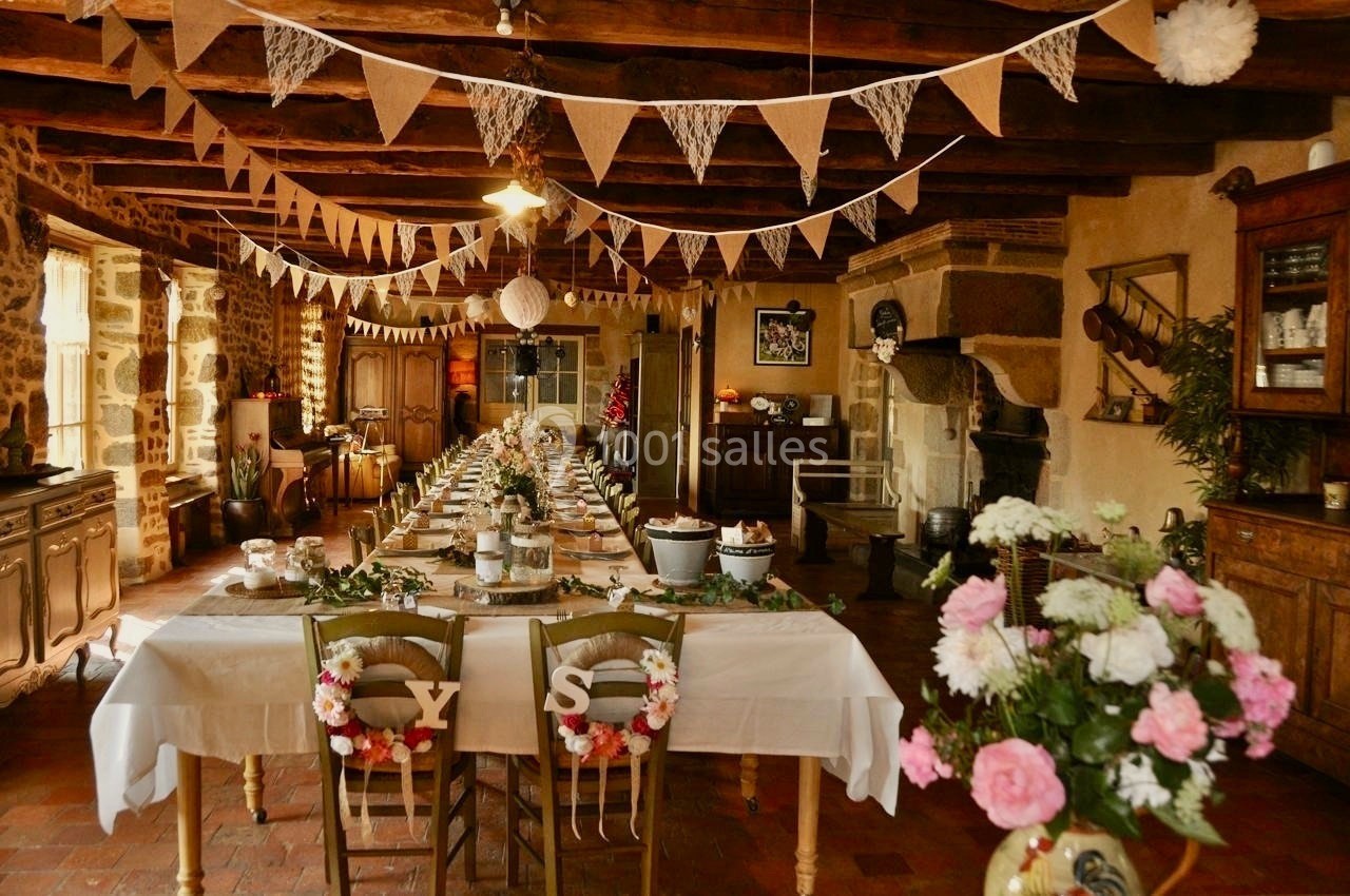 Salle rustique décorée pour un événement, avec longues tables dressées, guirlandes suspendues et fleurs en centre de table.