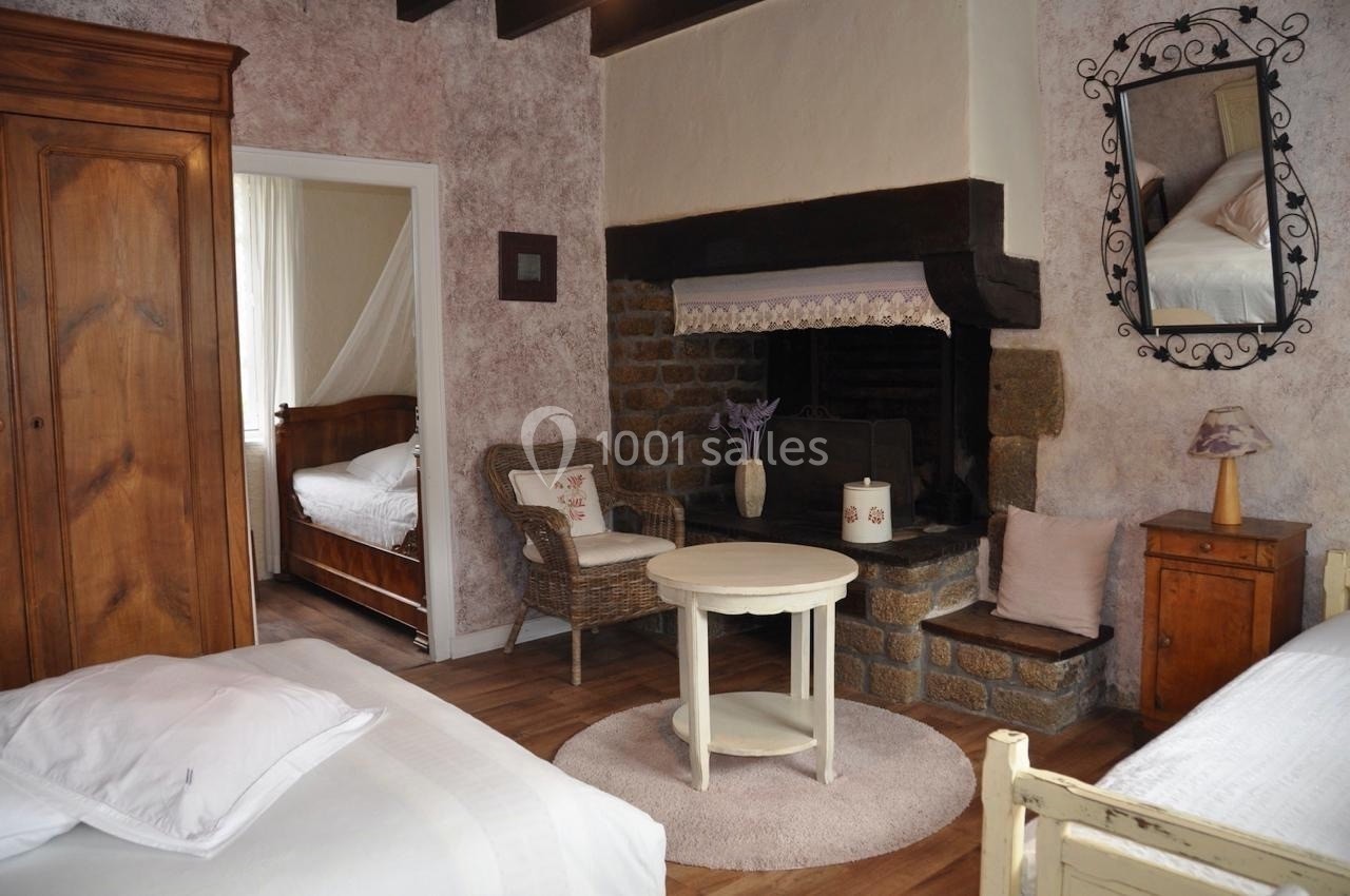 Chambre rustique avec lits, cheminée en pierre, armoire en bois, fauteuil en osier et table ronde au centre.