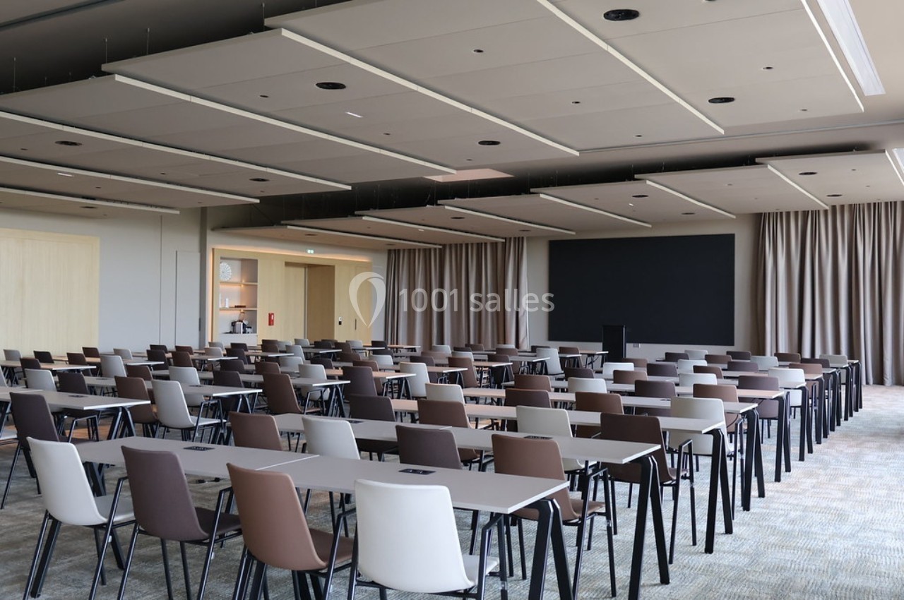 Salle de conférence moderne avec rangées de tables et chaises alignées, écran noir et rideaux en arrière-plan.