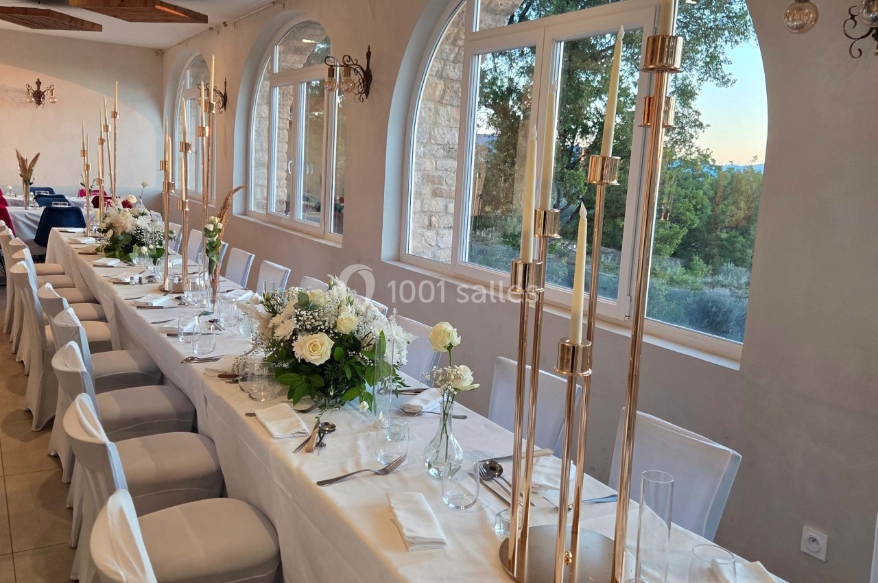 Grande table de réception décorée avec des fleurs blanches, des chandeliers dorés et une vue sur un paysage extérieur…