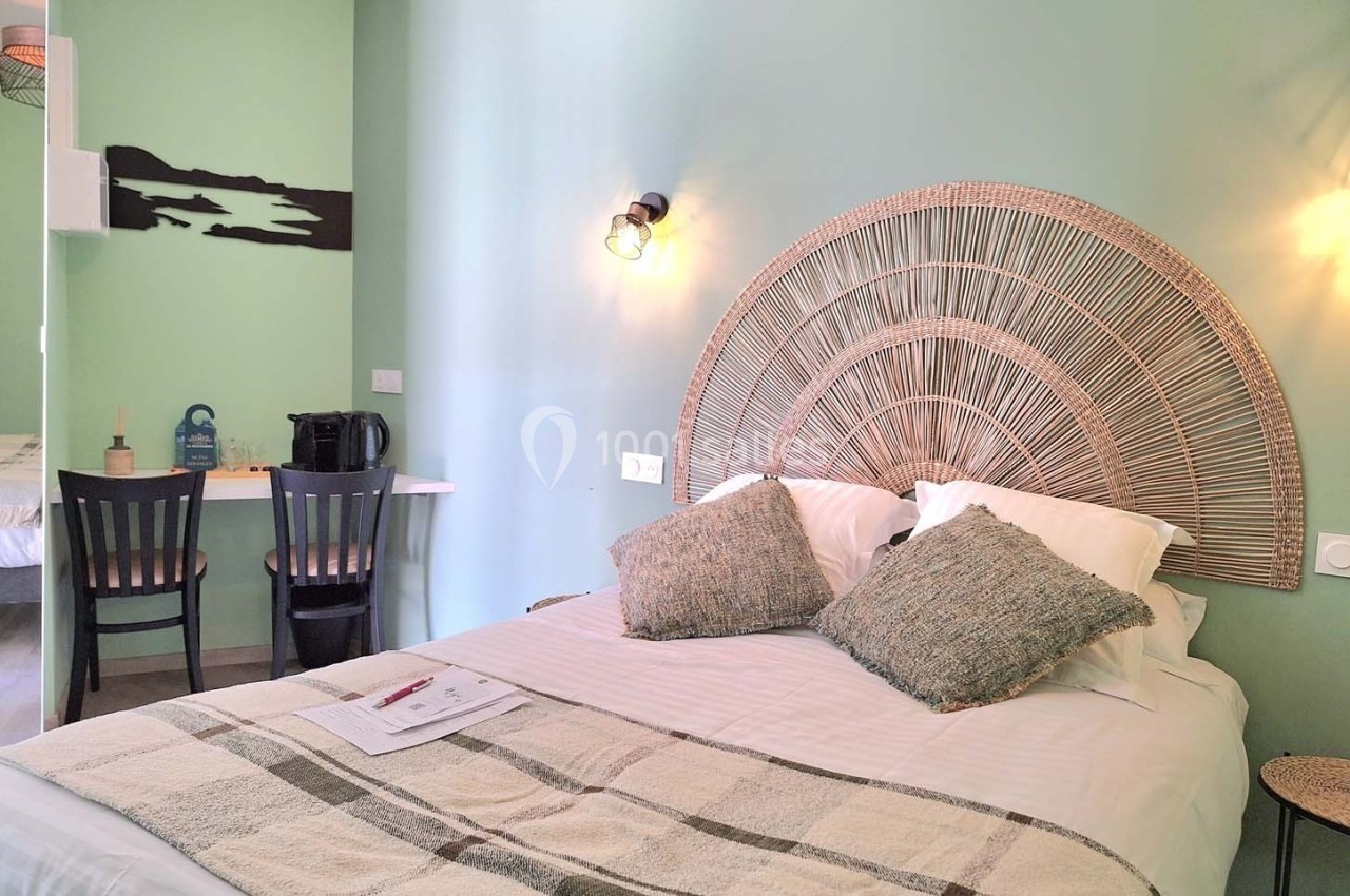 Chambre avec lit double, tête de lit en rotin, mur vert pastel, bureau avec chaises et plateau de courtoisie.