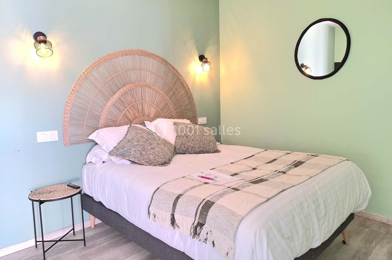 Lit double avec tête de lit en rotin, couvertures et coussins, dans une chambre aux murs vert clair avec éclairage mural.