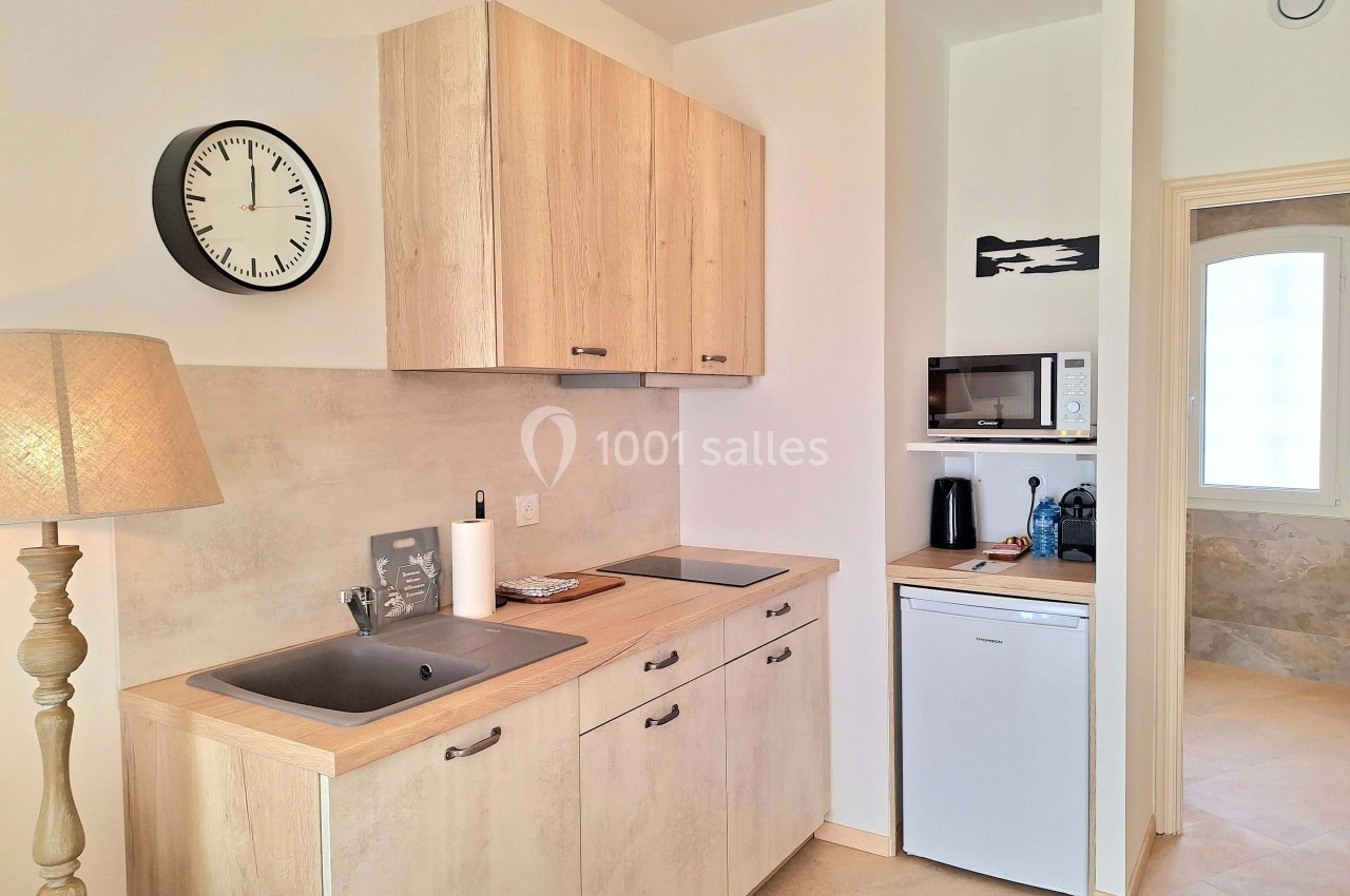 Cuisine compacte avec évier, plaques de cuisson, micro-ondes, réfrigérateur, horloge murale et lampe sur pied.