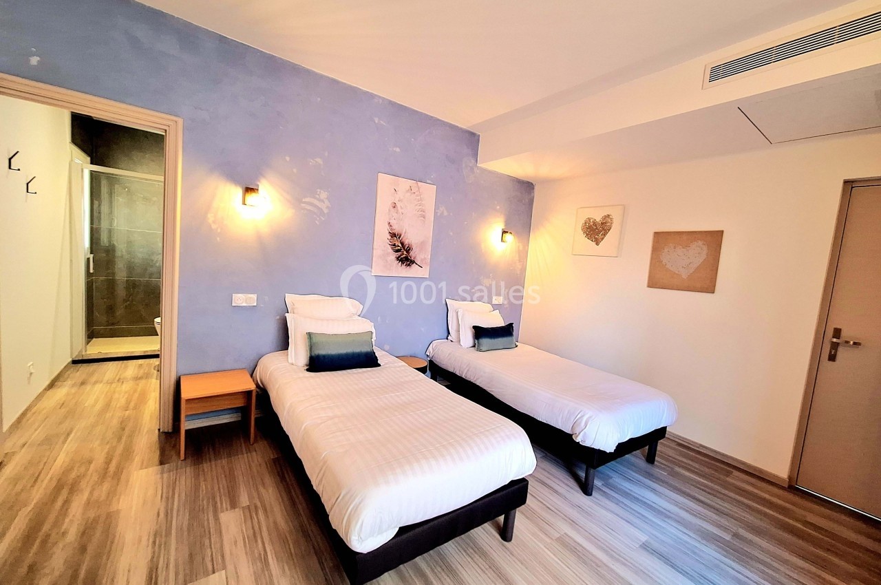 Chambre lumineuse avec deux lits simples, mur bleu, parquet clair et salle de bain attenante visible à gauche.