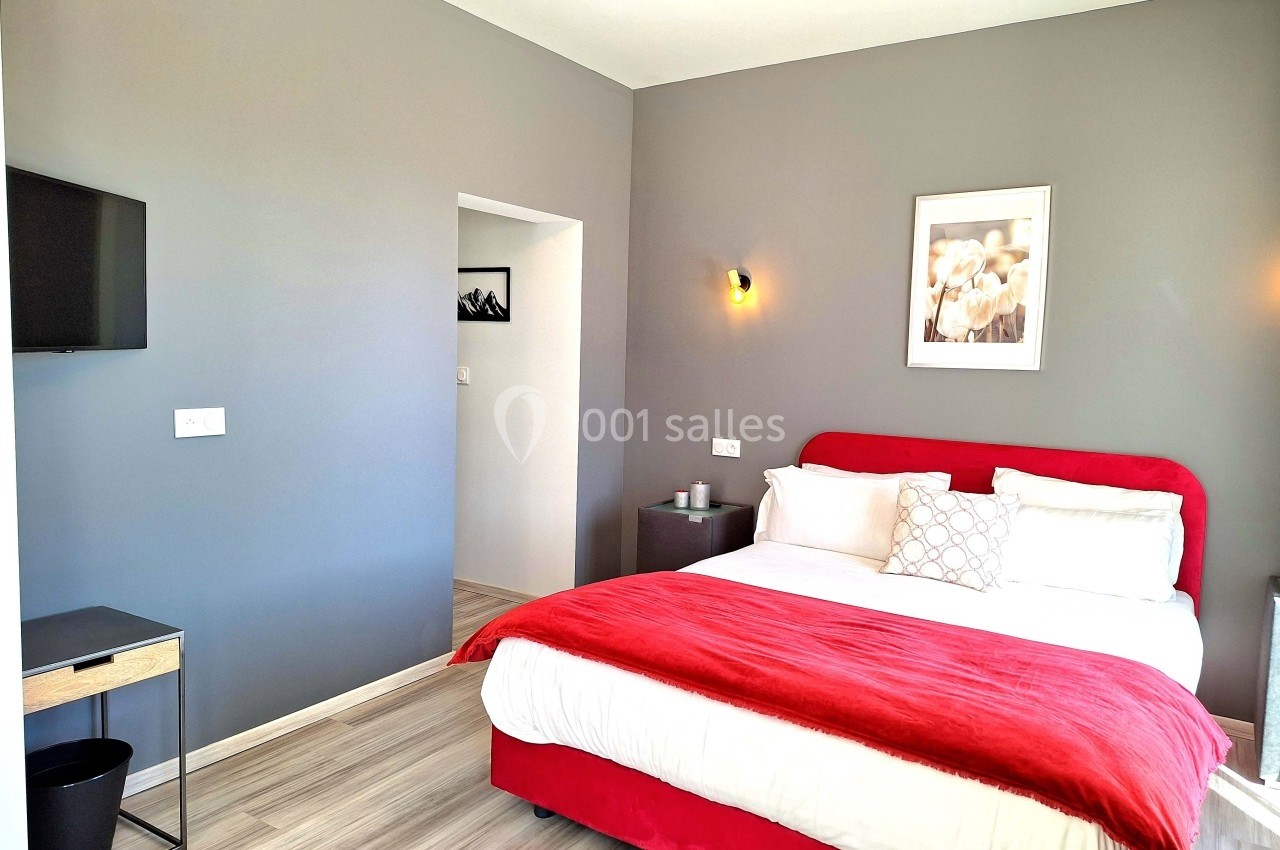 Chambre moderne avec lit double, mur gris, linge rouge et blanc, table de chevet, télévision et sol en bois clair.