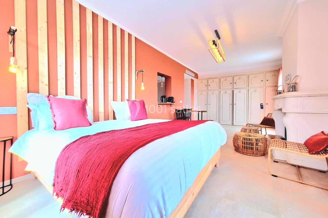 Chambre lumineuse avec lit double, mur en bois, coussins rouges, cheminée blanche et mobilier en osier.