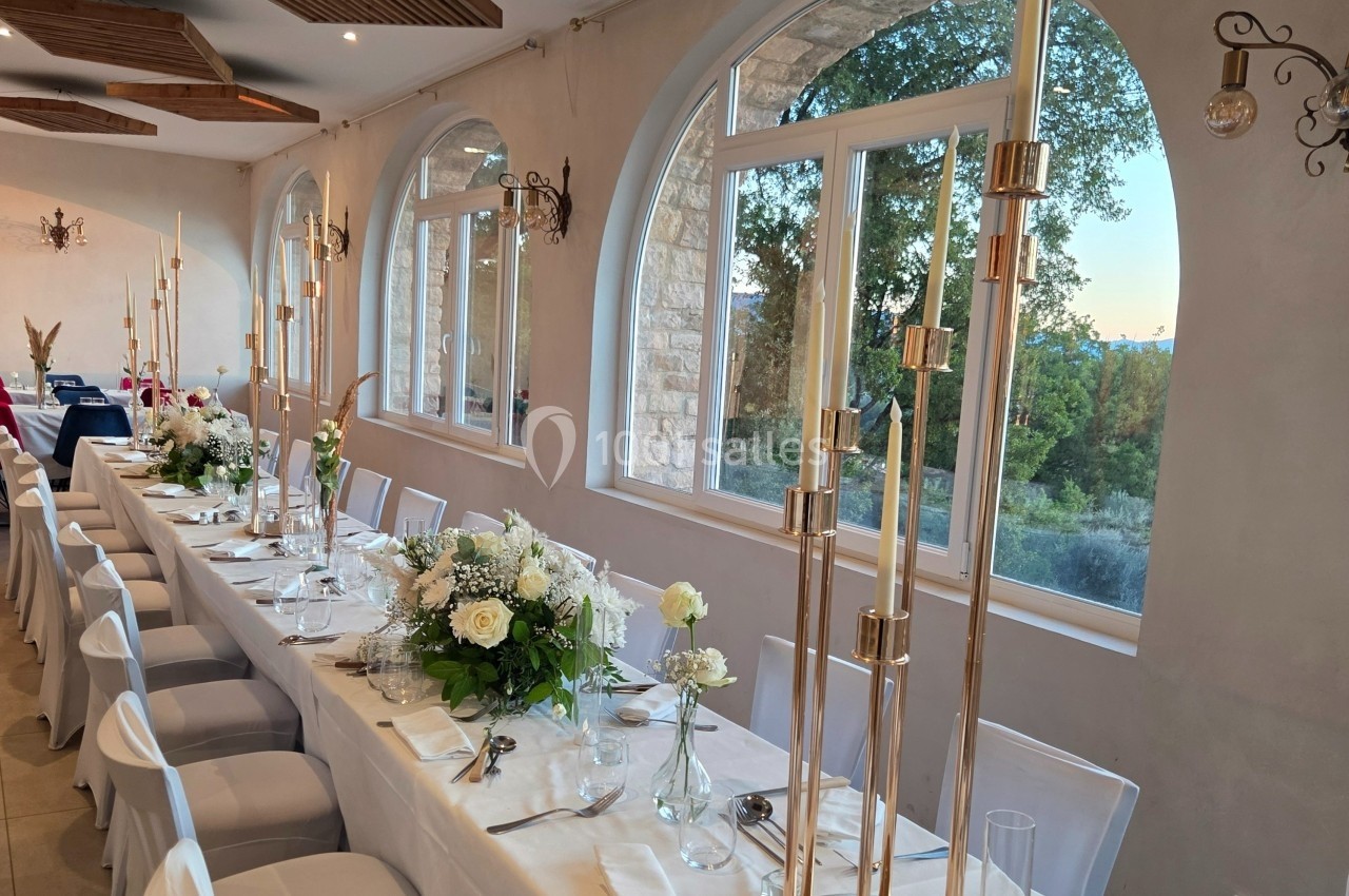 Salle de réception lumineuse avec une longue table décorée de fleurs blanches et de chandeliers dorés, vue sur l'extérieur.
