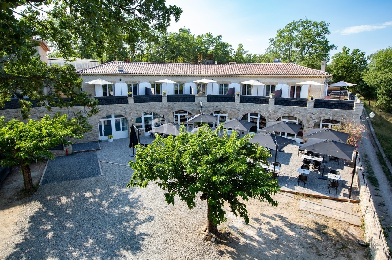 Bâtiment en pierre à deux étages avec terrasse ombragée par des parasols, entouré d'arbres et d'un espace gravillonné.