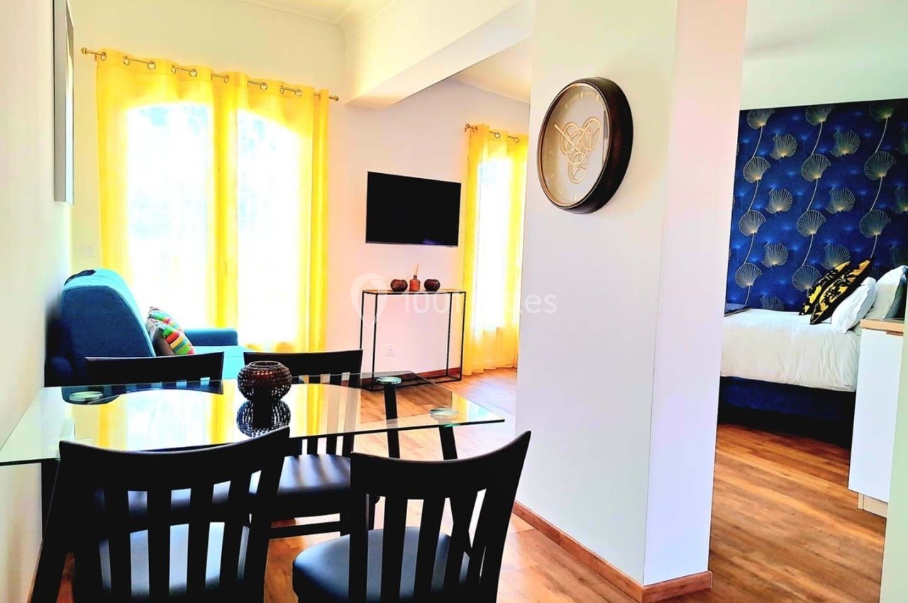 Salle à manger avec table en verre et chaises noires, salon lumineux avec rideaux jaunes, et chambre adjacente.