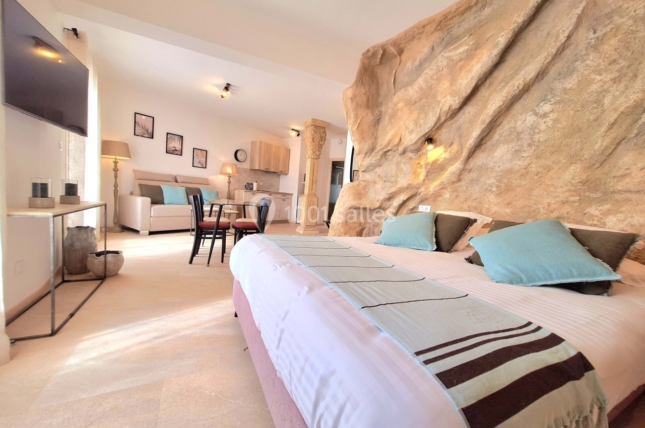 Chambre spacieuse avec lit double, coin salon, cuisine ouverte et mur en pierre naturelle éclairé.