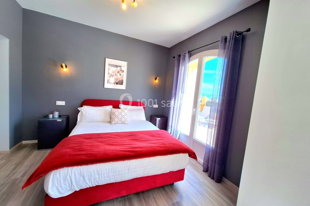 Chambre lumineuse avec lit double, murs gris, rideaux violets et grande fenêtre donnant sur une terrasse ensoleillée.