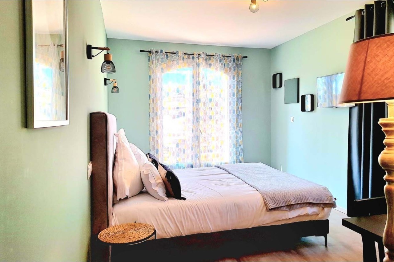 Chambre lumineuse avec murs verts, lit double, lampes murales, rideaux à motifs et mobilier en bois.