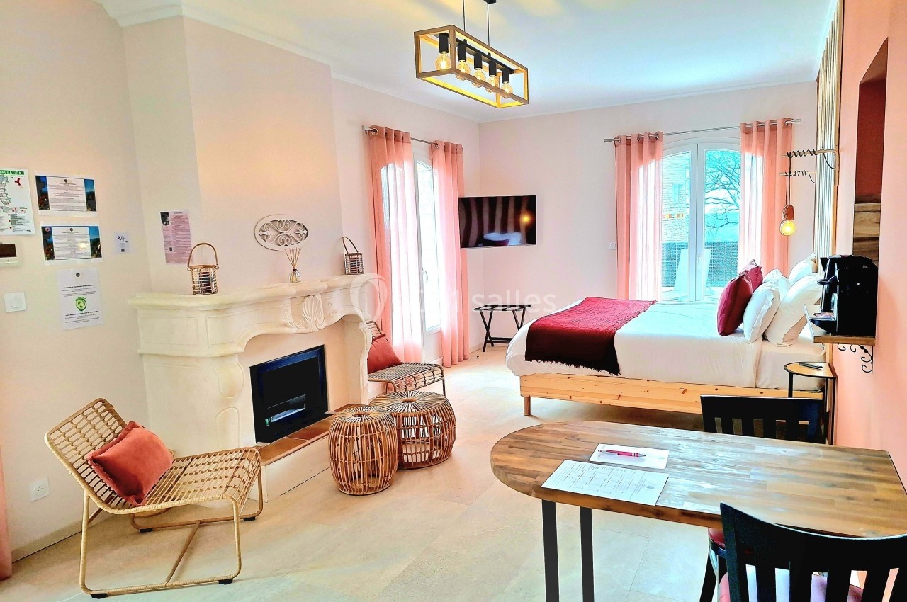 Chambre lumineuse avec lit double, cheminée décorative, coin salon, bureau en bois et grandes fenêtres avec rideaux roses.