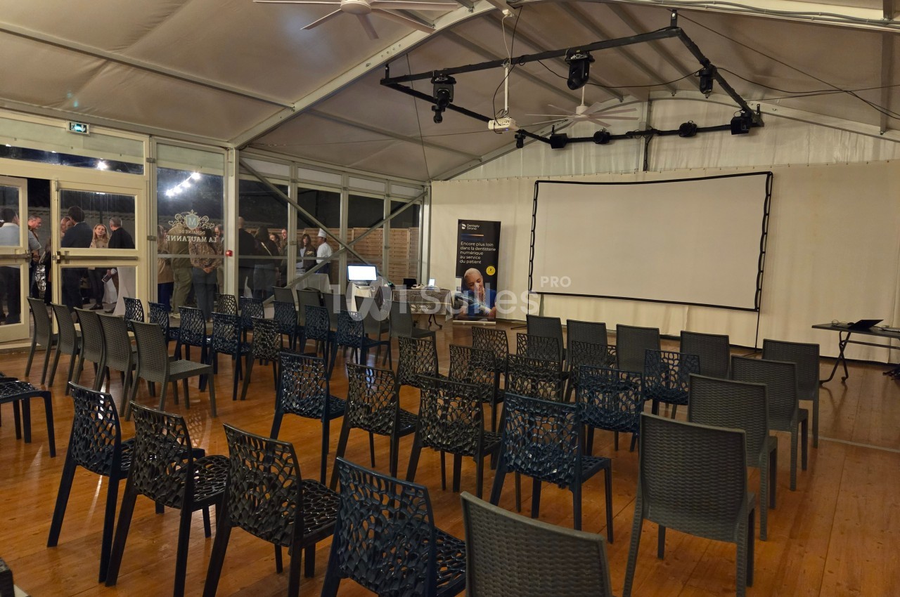 Salle de conférence vide avec des rangées de chaises en plastique, un écran de projection et un éclairage suspendu.