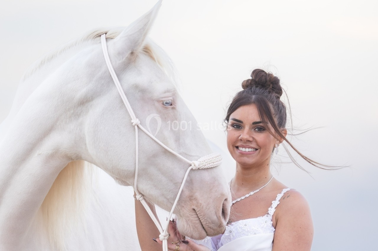 Femme souriante en robe blanche tenant un cheval blanc avec un licol, sur un fond clair et flou.