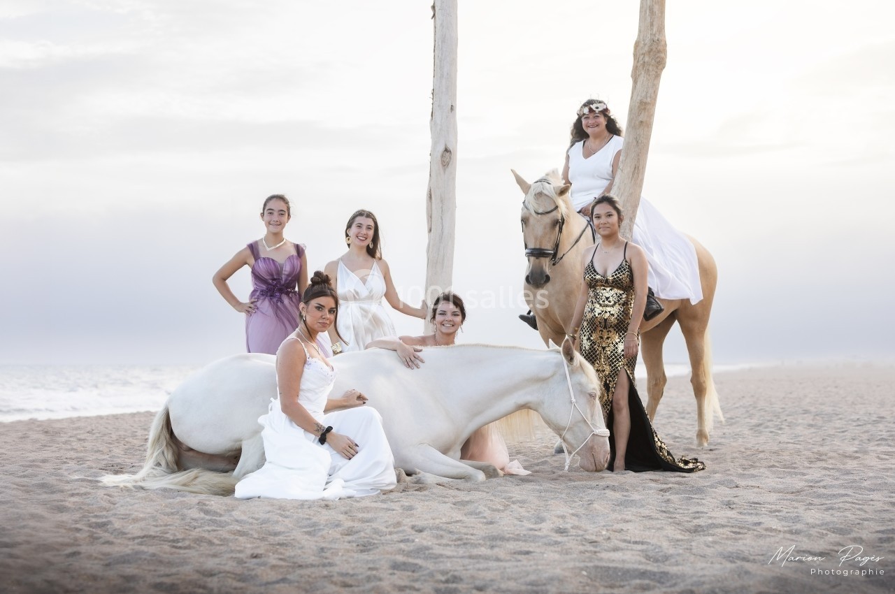 Cinq femmes en robes élégantes posent avec deux chevaux sur une plage au coucher du soleil.