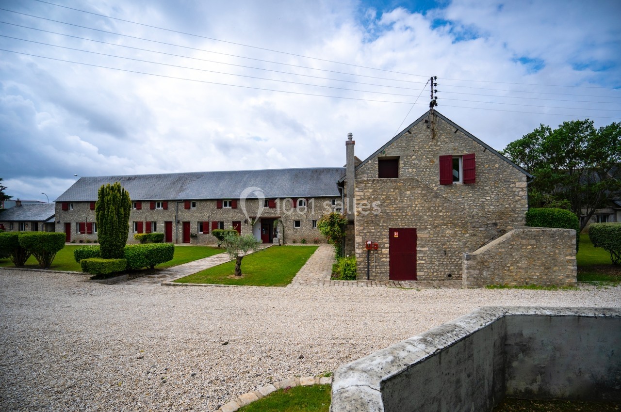 Bâtiment en pierre avec volets rouges, entouré de pelouses et d'arbustes taillés, sous un ciel nuageux.