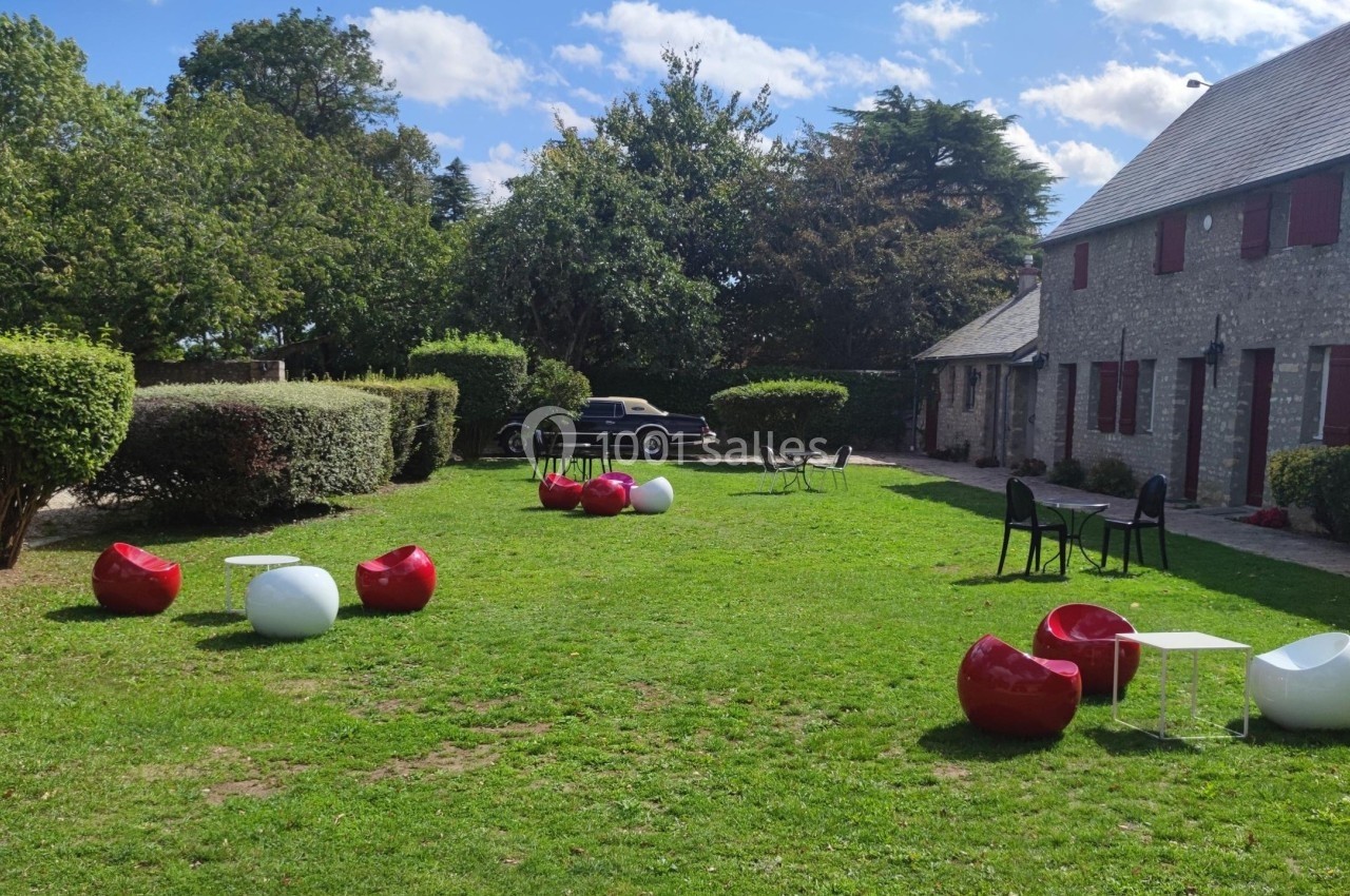 Espace extérieur avec pelouse, mobilier design rouge et blanc, tables basses, et bâtiment en pierre à l'arrière-plan.