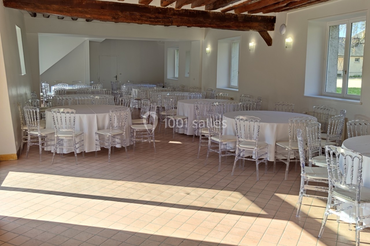 Salle lumineuse avec tables rondes dressées de nappes blanches et chaises transparentes, poutres apparentes au plafond.