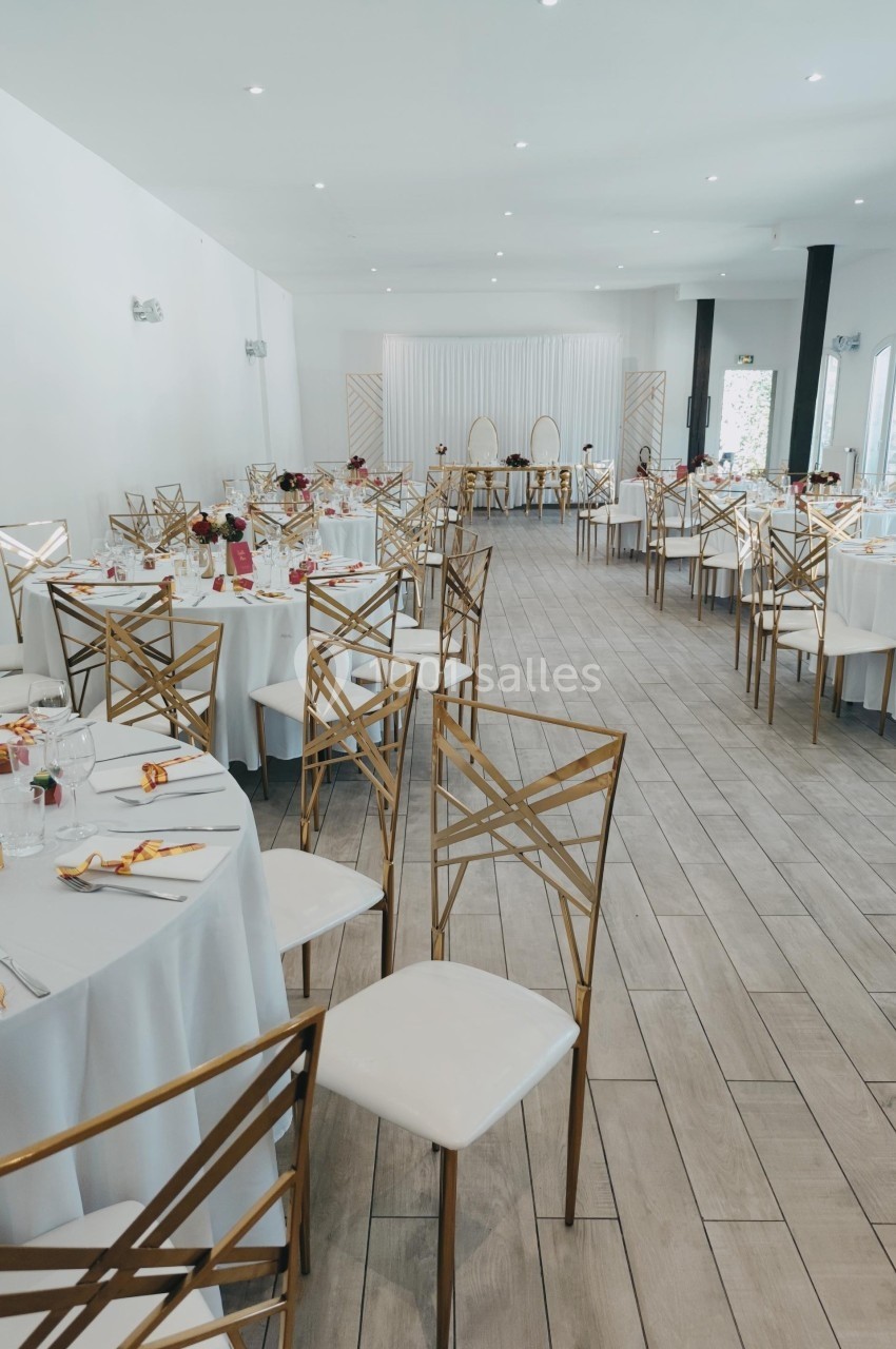 Salle de réception lumineuse avec tables rondes dressées, chaises dorées et décorations élégantes.