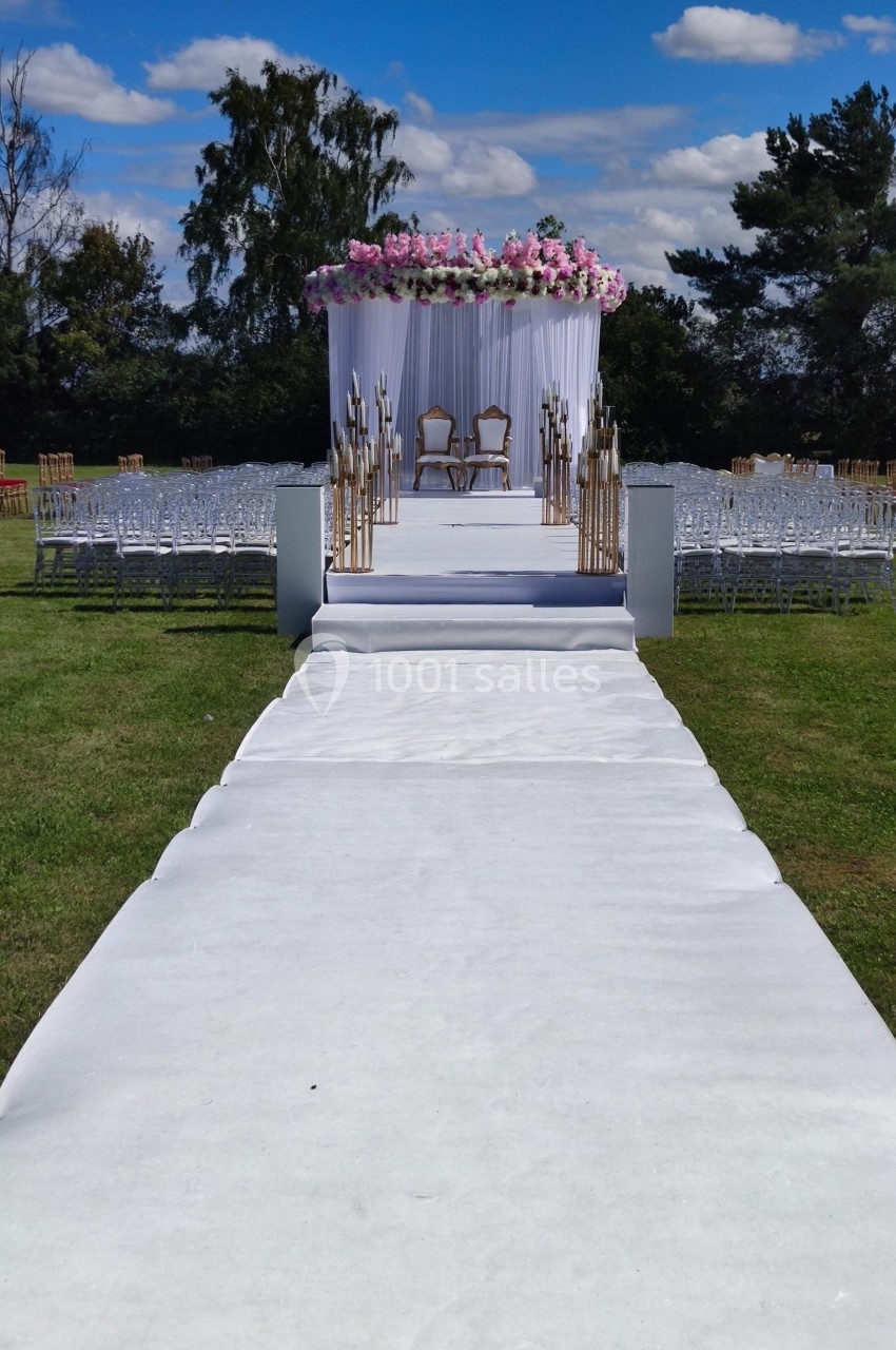 Allée blanche menant à une arche décorée de fleurs roses et blanches avec deux chaises, en extérieur sur pelouse.