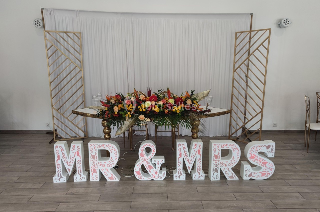 Décoration de mariage avec des lettres ’MR & MRS’, une table ornée de fleurs colorées et un fond blanc élégant.