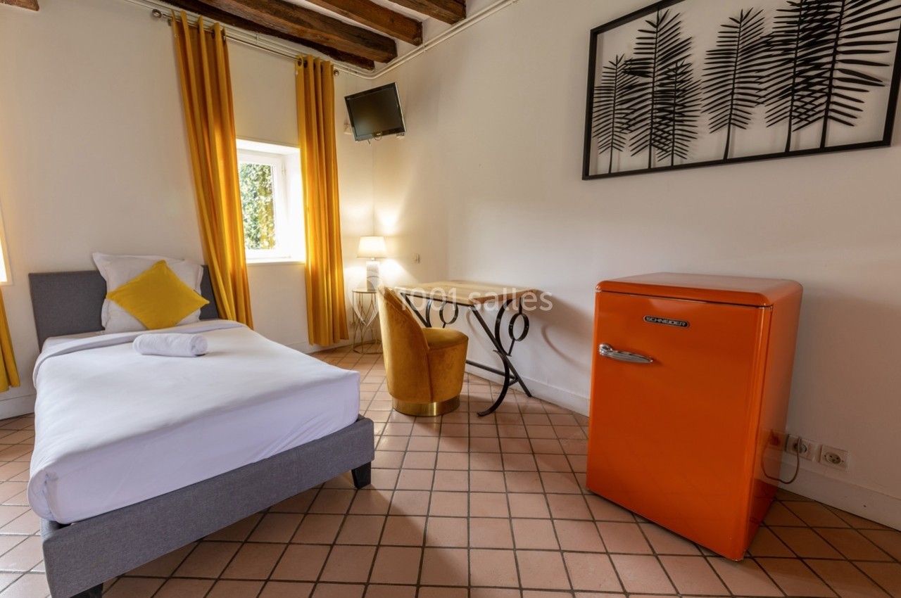 Chambre lumineuse avec lit simple, réfrigérateur orange, bureau en bois, fauteuil jaune et rideaux assortis.