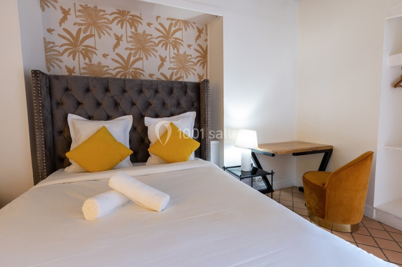 Lit double avec coussins jaunes, serviettes roulées, bureau en bois, chaise jaune et mur décoré de motifs tropicaux.